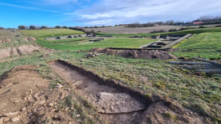 Photographie en couleur du site archéologique d’Aquae Segetae, en cours de fouille. Vue générale des vestiges en terre et pierre, avec des tranchées de fouille, des murs partiellement dégagés parmi l'herbe. En arrière-plan, un paysage de reliefs verdoyants. Image réalisée par J. Spiesser, CD45